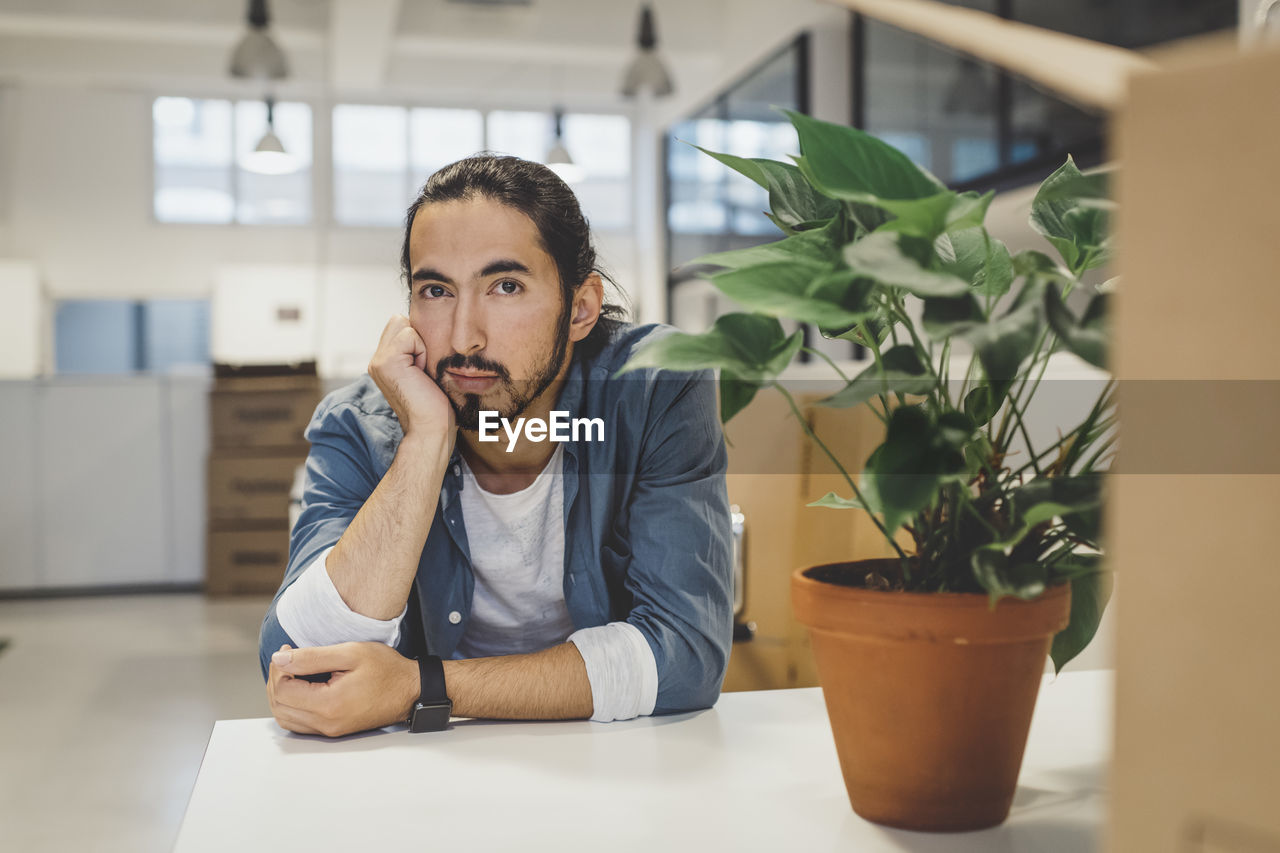 Portrait of young businessman with hand on chin sitting at table in new office