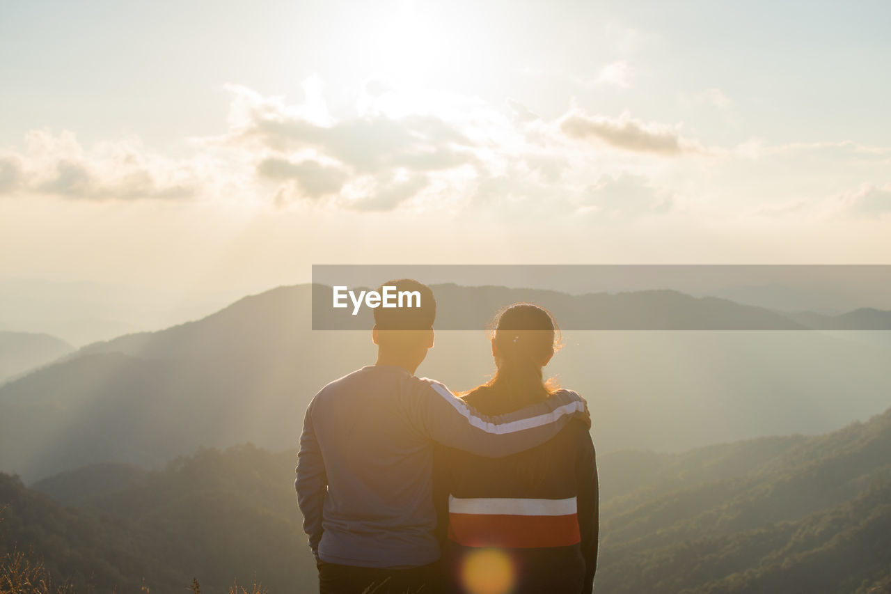Rear view of couple standing on mountain against sky during sunset