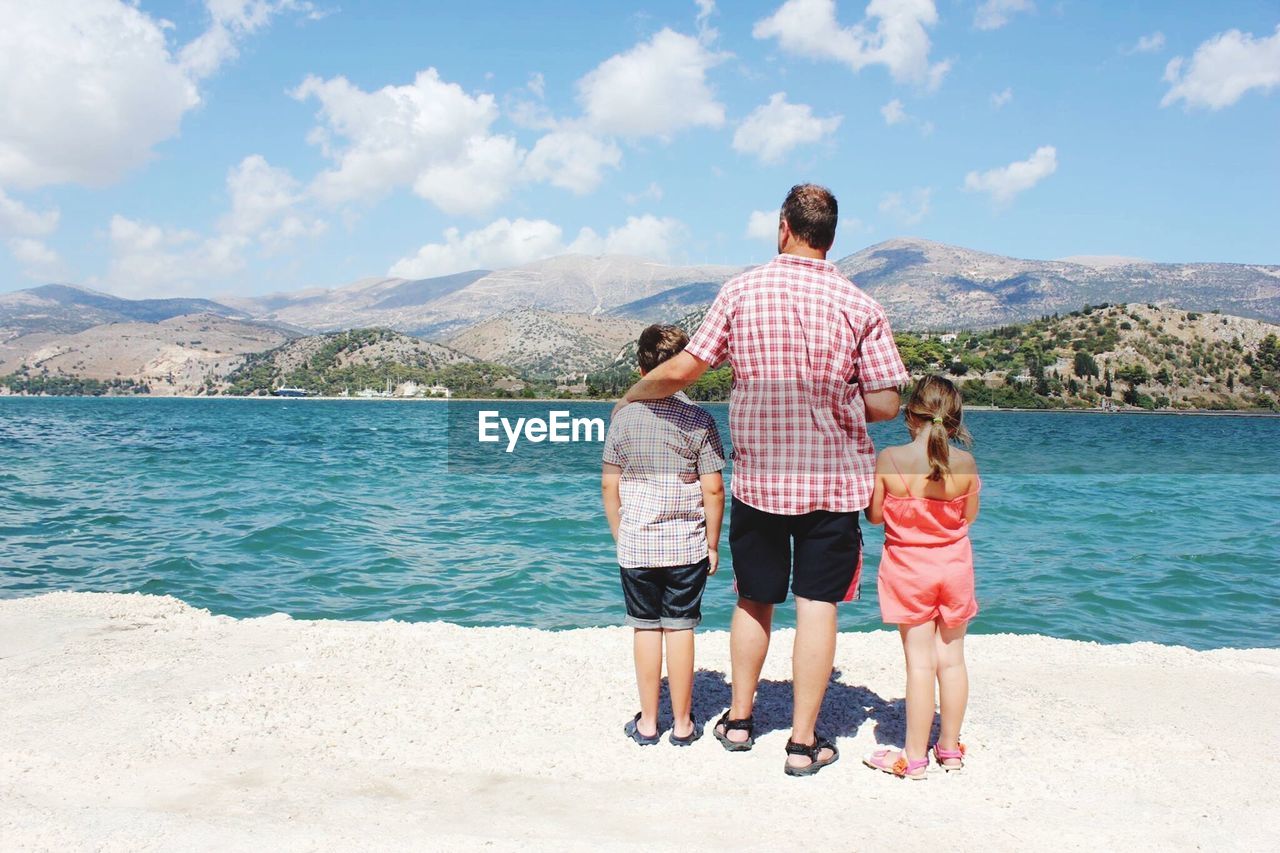 Rear view of father with children standing on shore at beach against sky