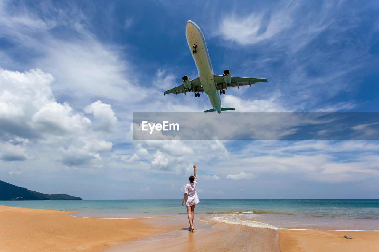 Low angle view of airplane flying over woman at beach against sky