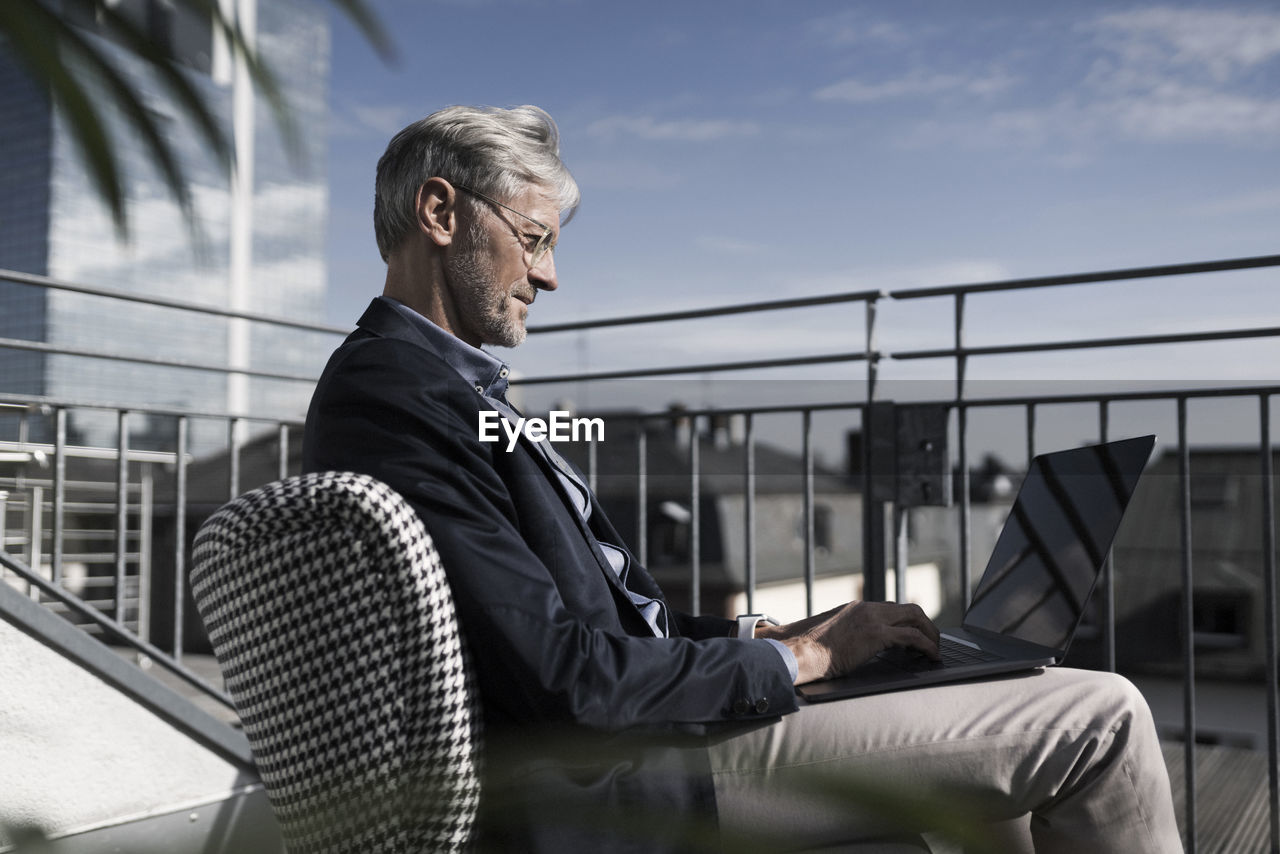 Grey-haired businessman sitting on balcony using laptop