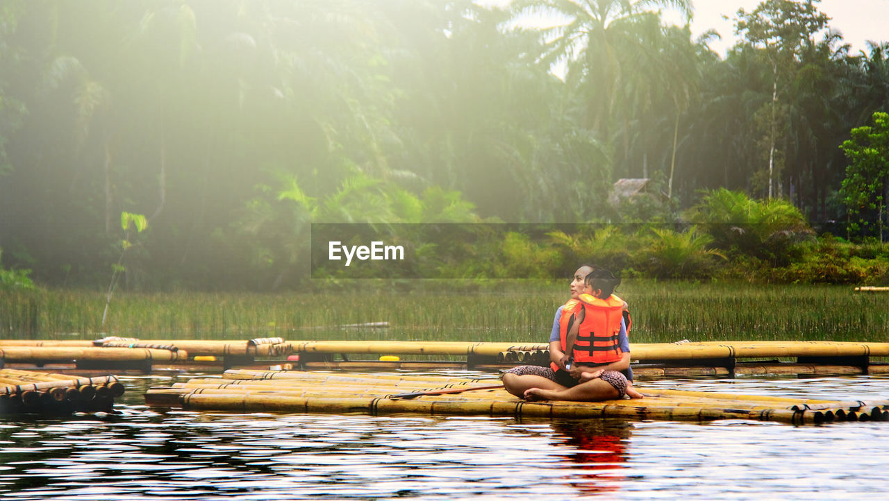 Mother and daughter wearing life jackets sitting on wooden raft 