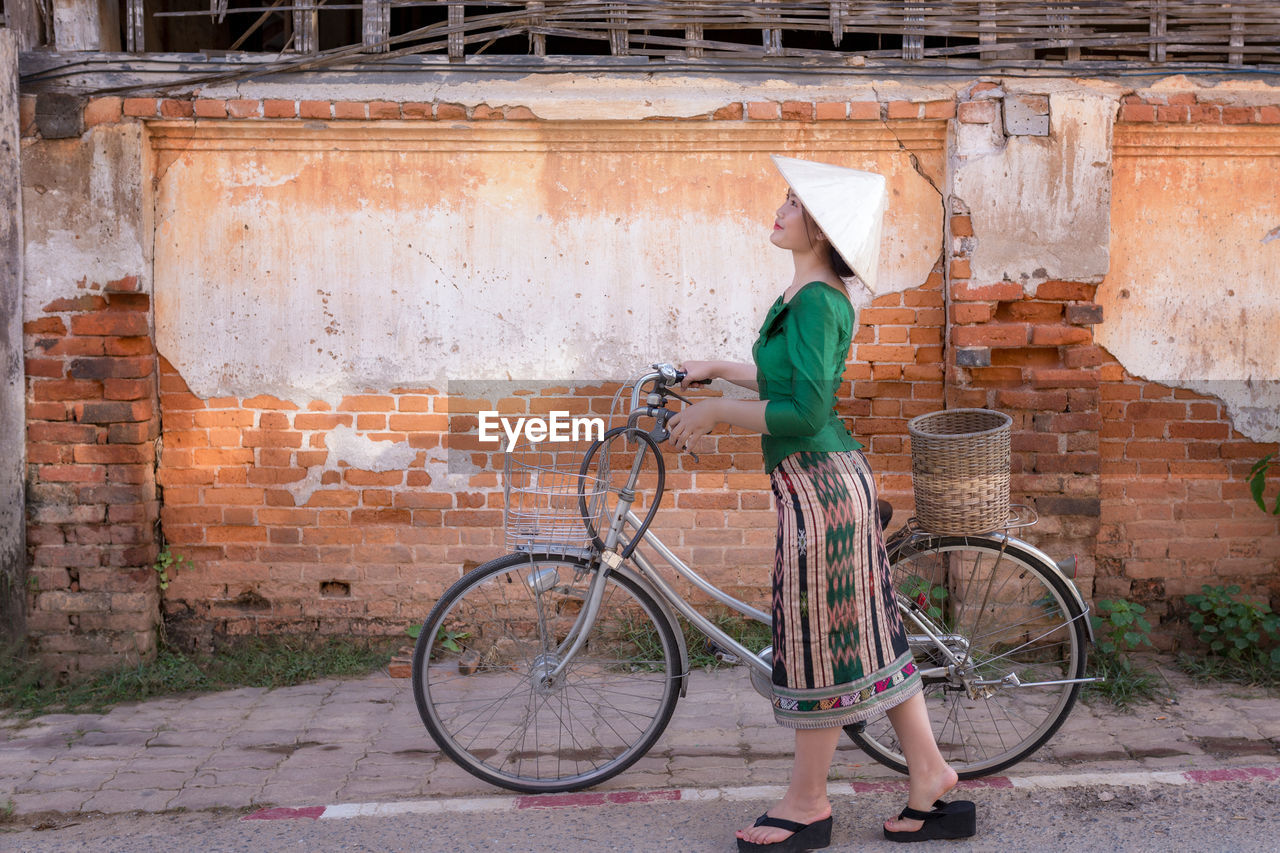 Side view of woman holding bicycle while walking on street against brick wall