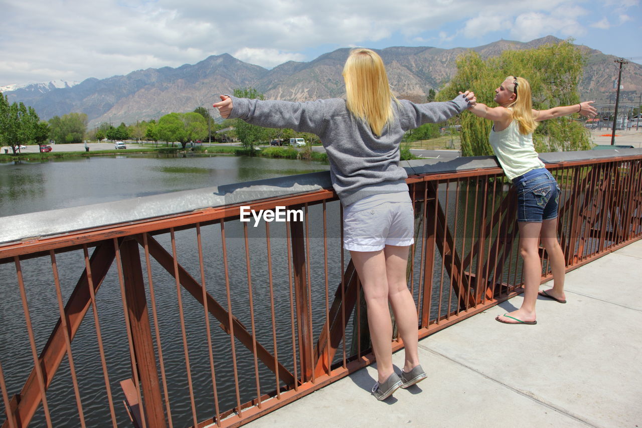 Teenage girls with arms outstretched standing on footbridge over river against mountains