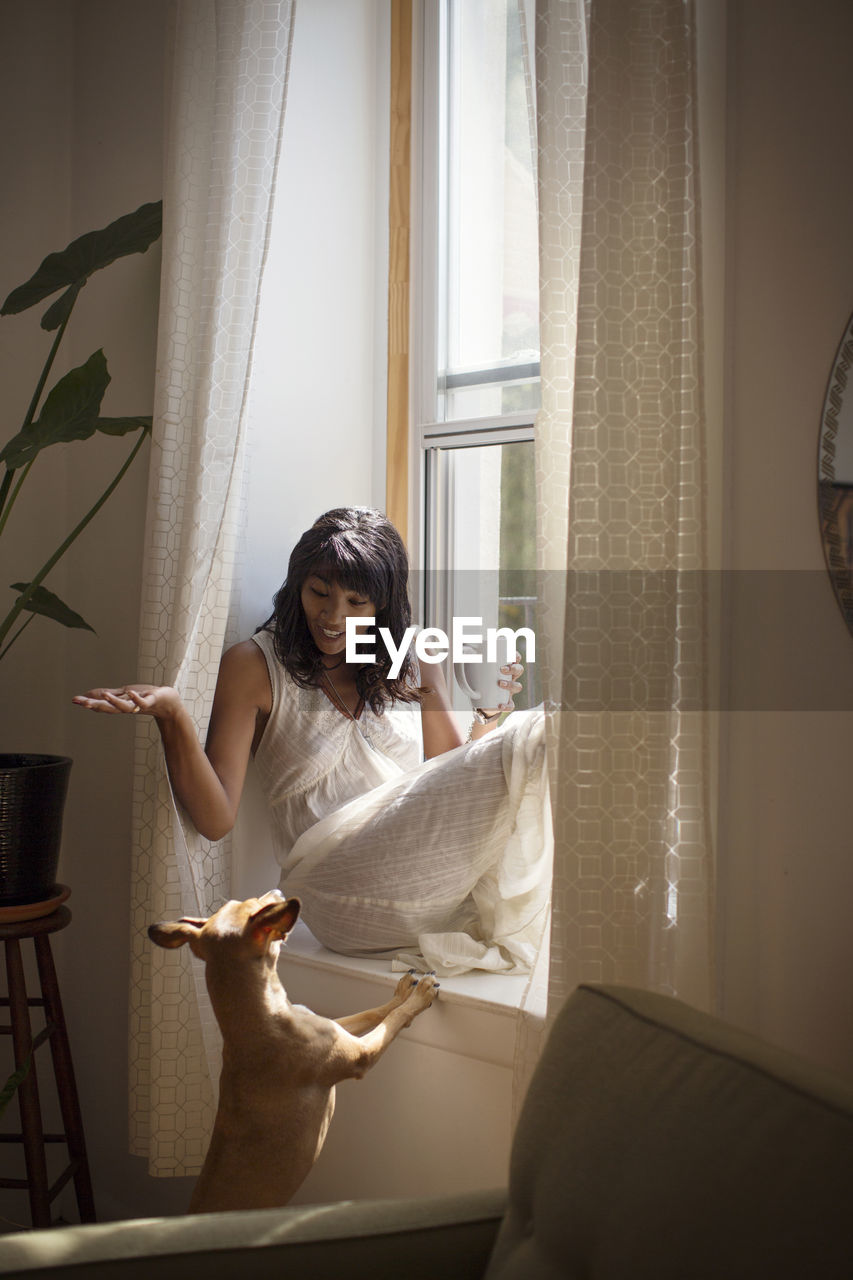 Woman looking at dog while sitting on window sill