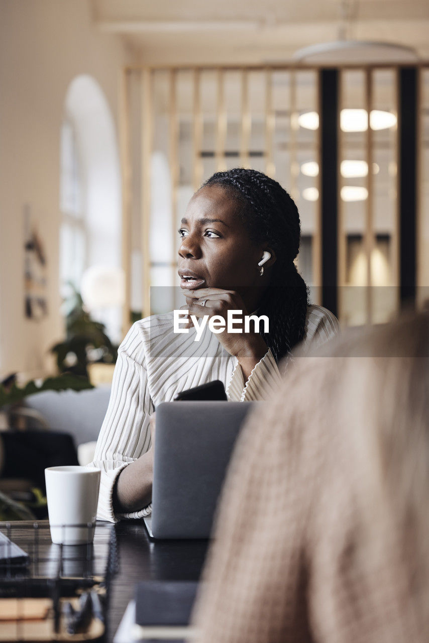 Thoughtful businesswoman looking away while talking through wireless in-ear headphones at coworking office