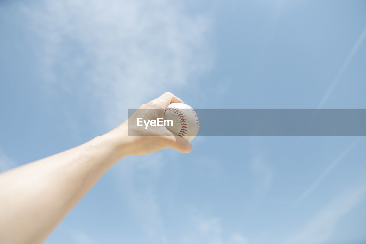 Cropped hand holding baseball against sky