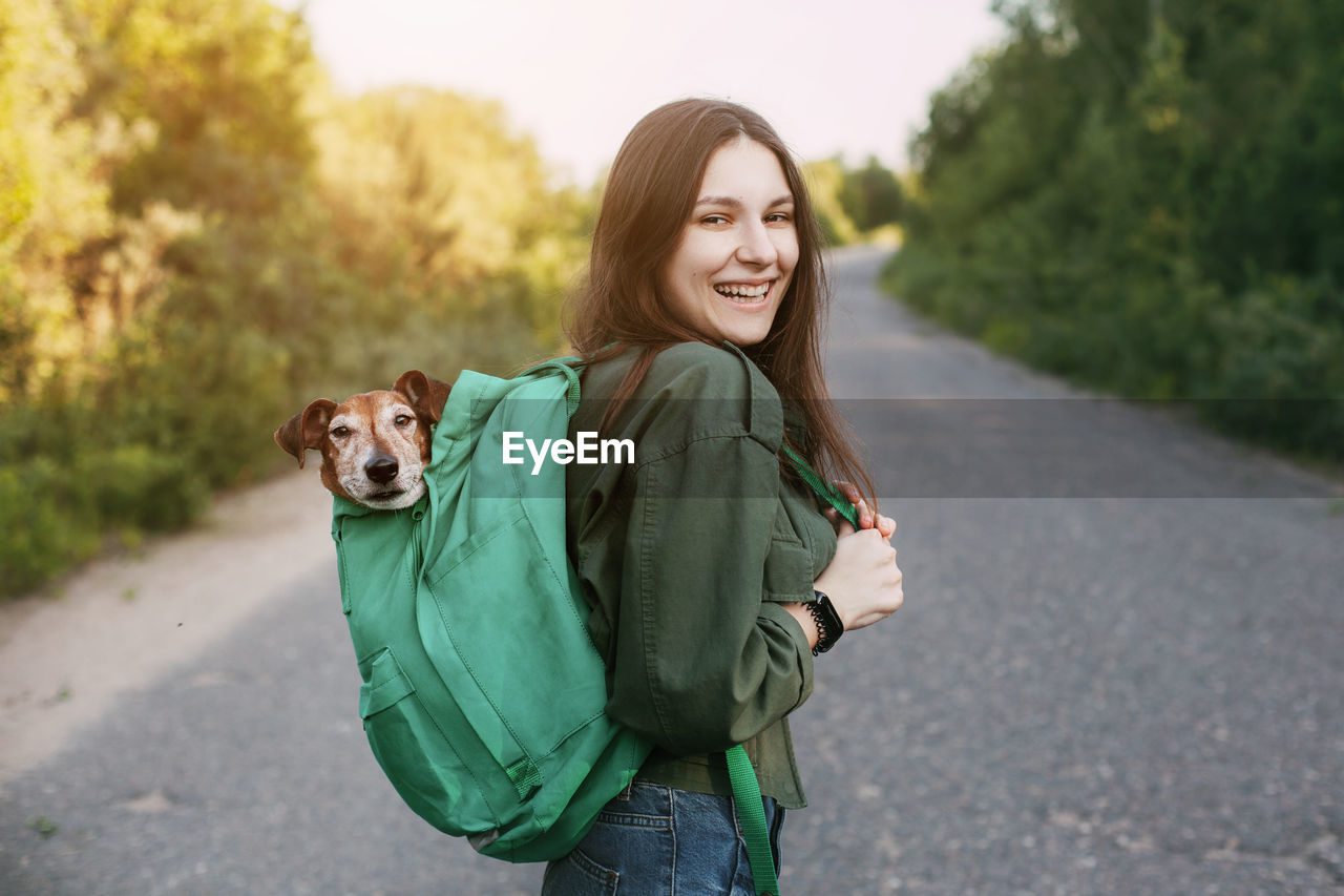 A smiling girl is holding a green backpack on her shoulder, from which a cute dog looks out