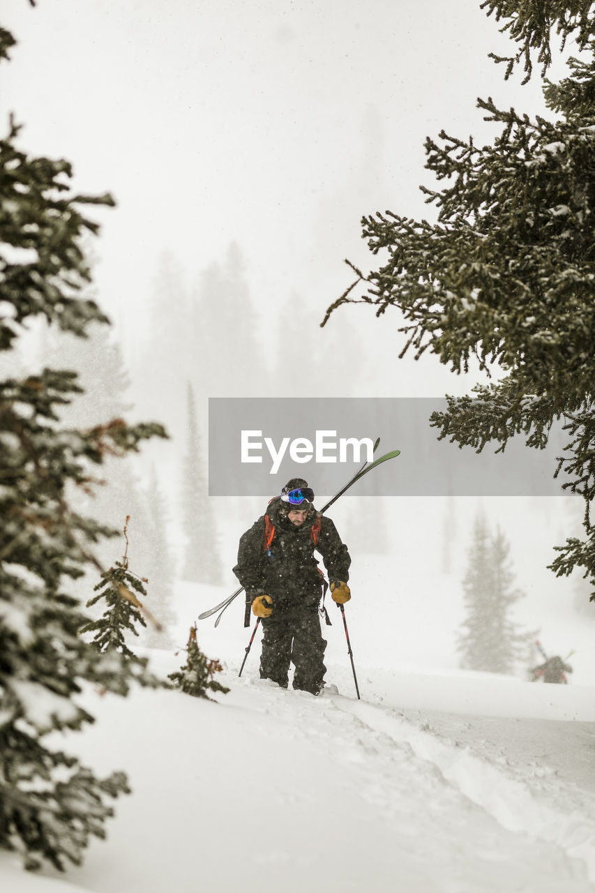 Man with skies walking on snow covered field in forest during snowfall