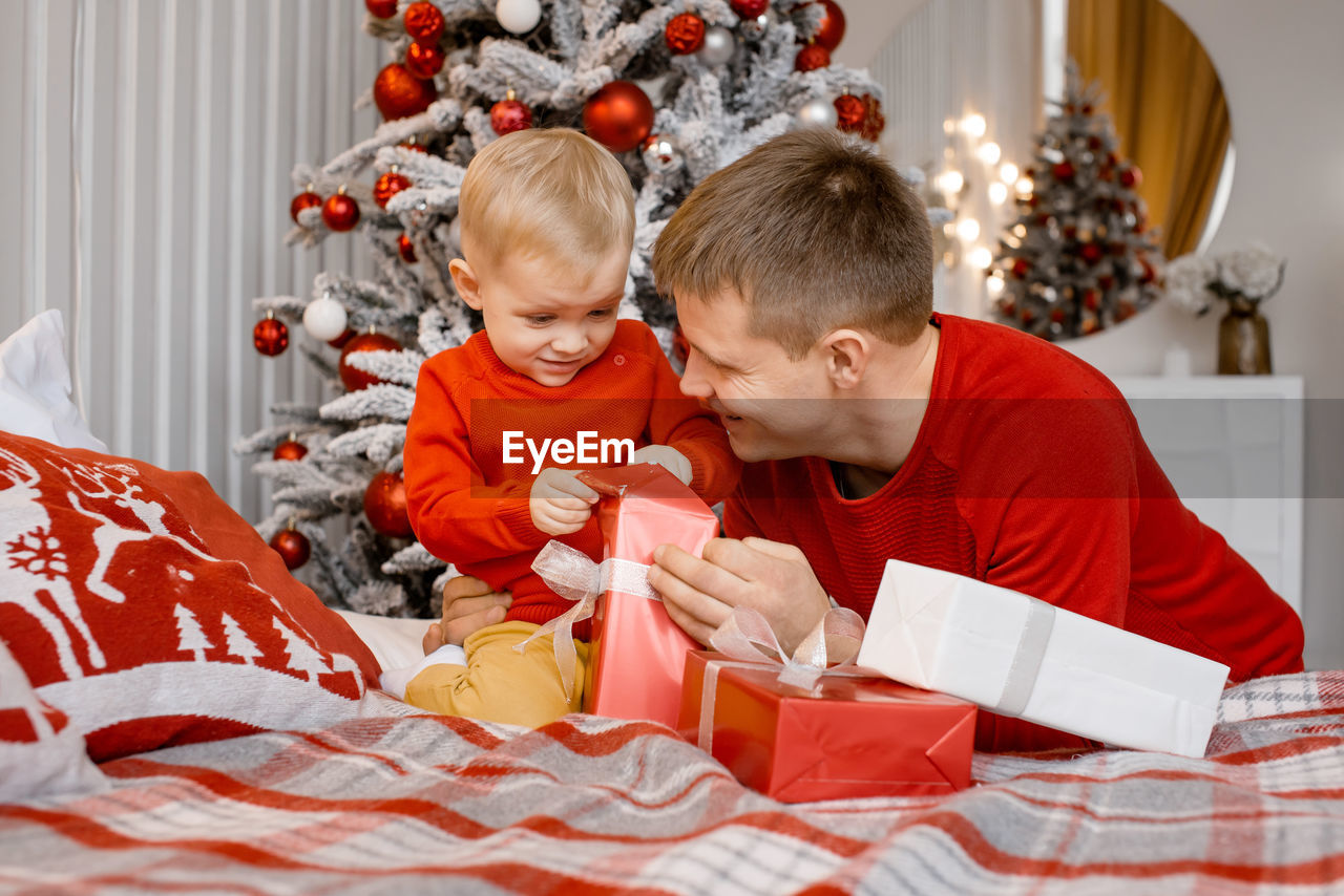 Happy smiling father and his little son unwrapping a present sitting on the bed near christmas tree