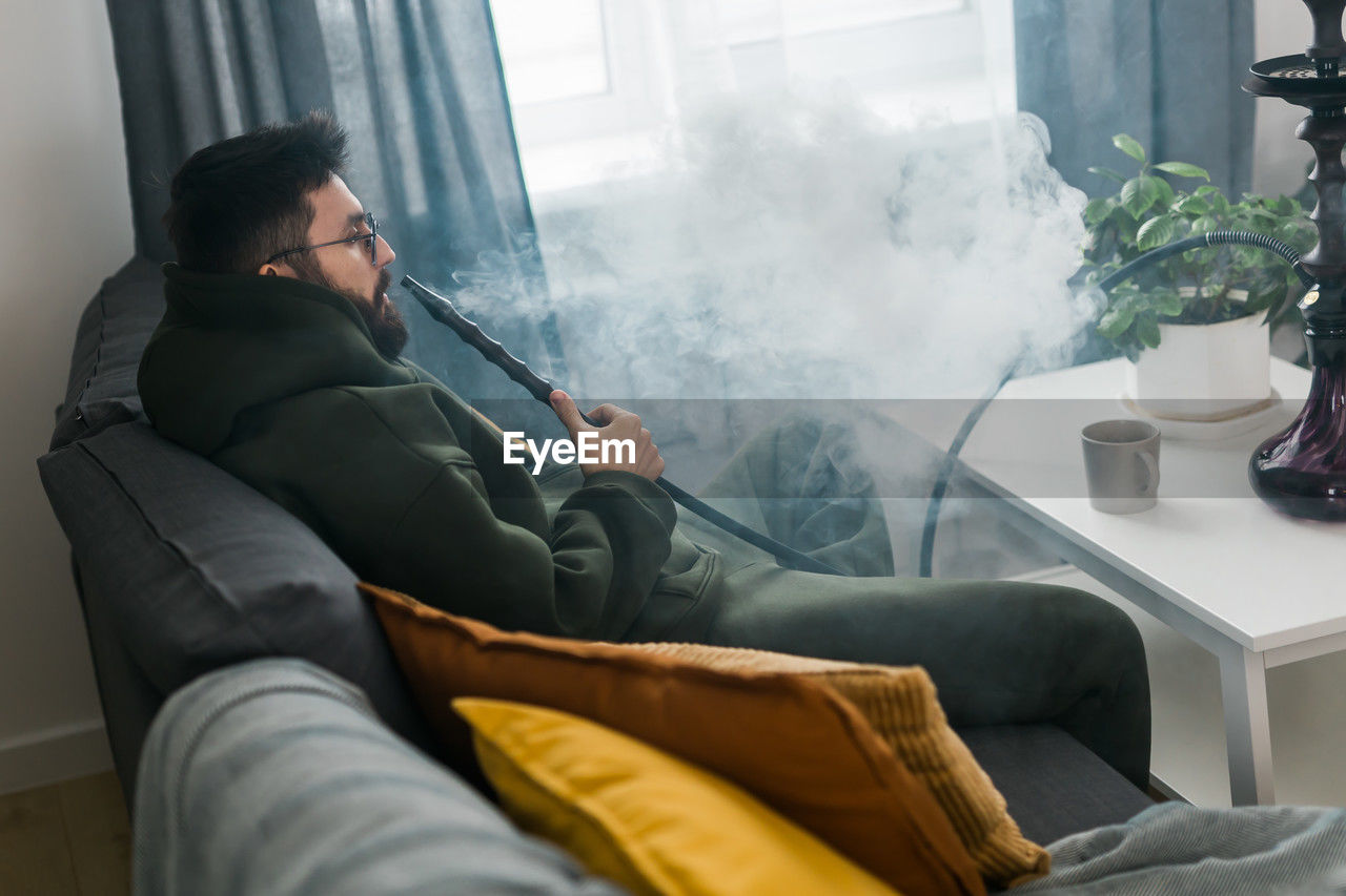 smoke, adult, one person, indoors, domestic room, men, furniture, window, communication, living room, person, domestic life, smoking, sofa, activity, sitting, lifestyles, relaxation, clothing, home interior, technology, casual clothing, side view, interior design, three quarter length, young adult, smoking issues, holding, nature, leisure activity, day
