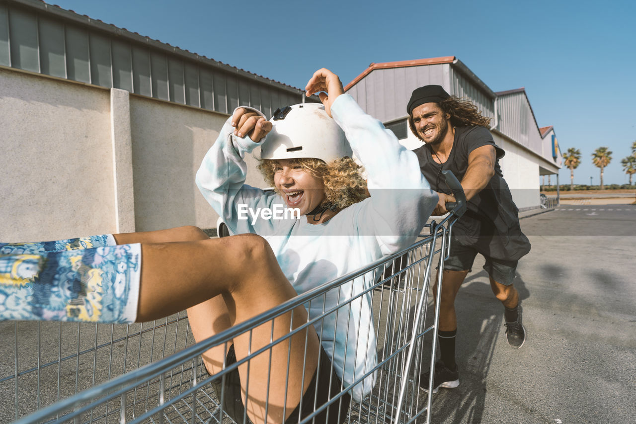 Playful young man pushing girlfriend sitting in shopping cart outside supermarket on sunny day