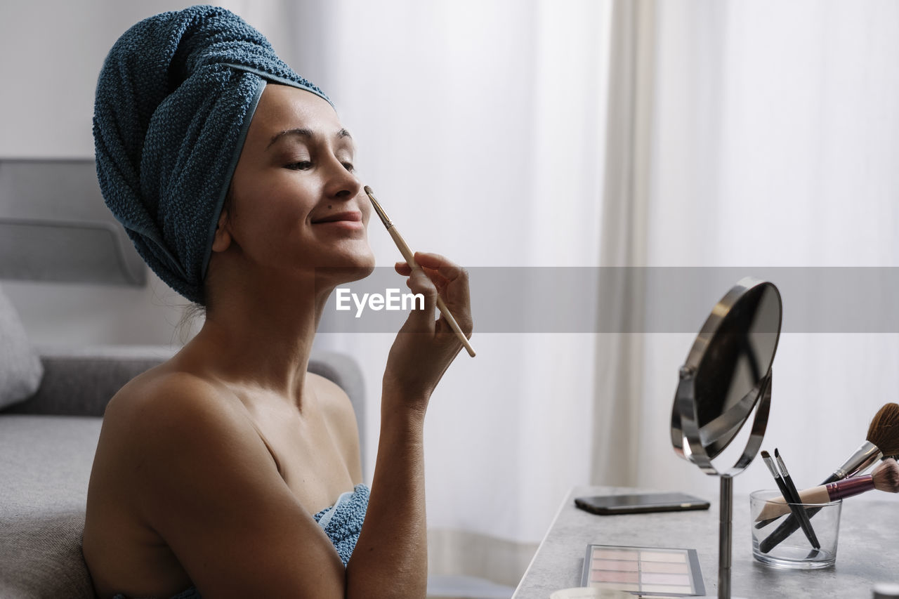 Content young bare shouldered female with towel on head applying eye shadow on eyebrows during makeup procedure after shower at home