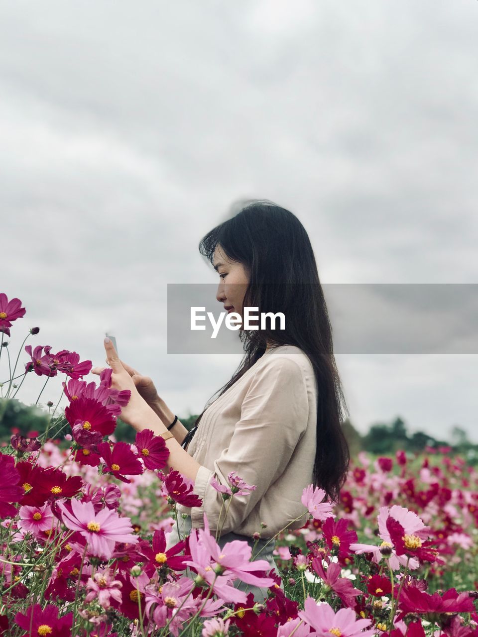 Side view of young woman photographing with mobile phone while standing on field against cloudy sky