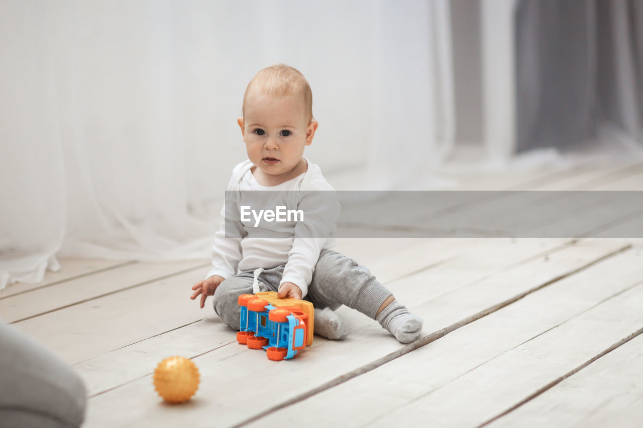 Baby of 10 months in light clothes sits on a wooden floor with bright toys, a light real interior.