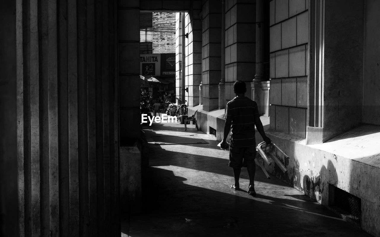 Rear view of man holding stool while walking on walkway by building