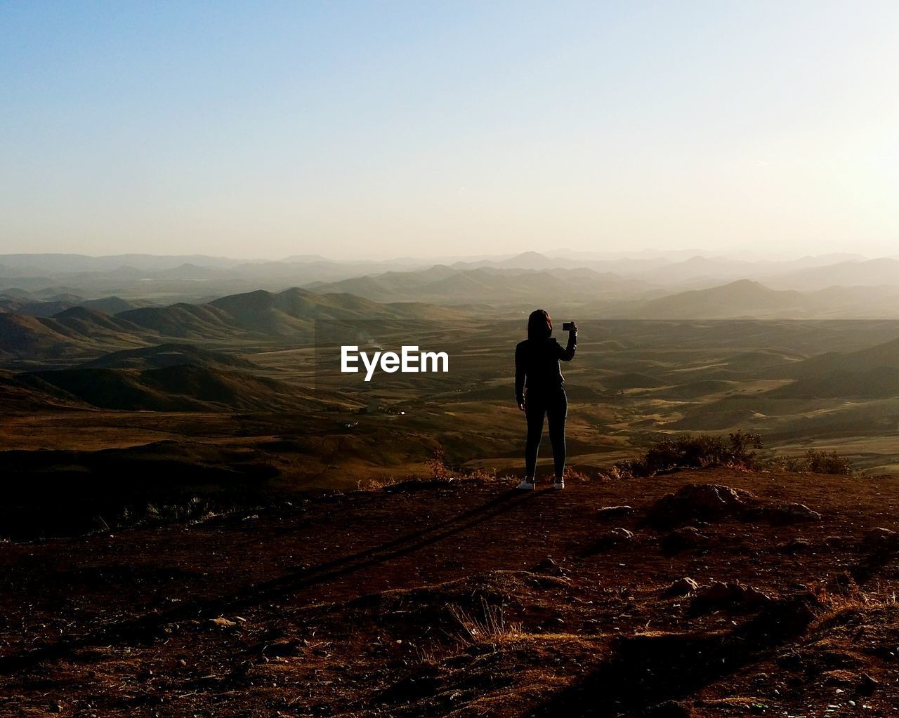 Rear view of silhouette mid adult woman photographing with mobile phone while standing on mountain against clear sky