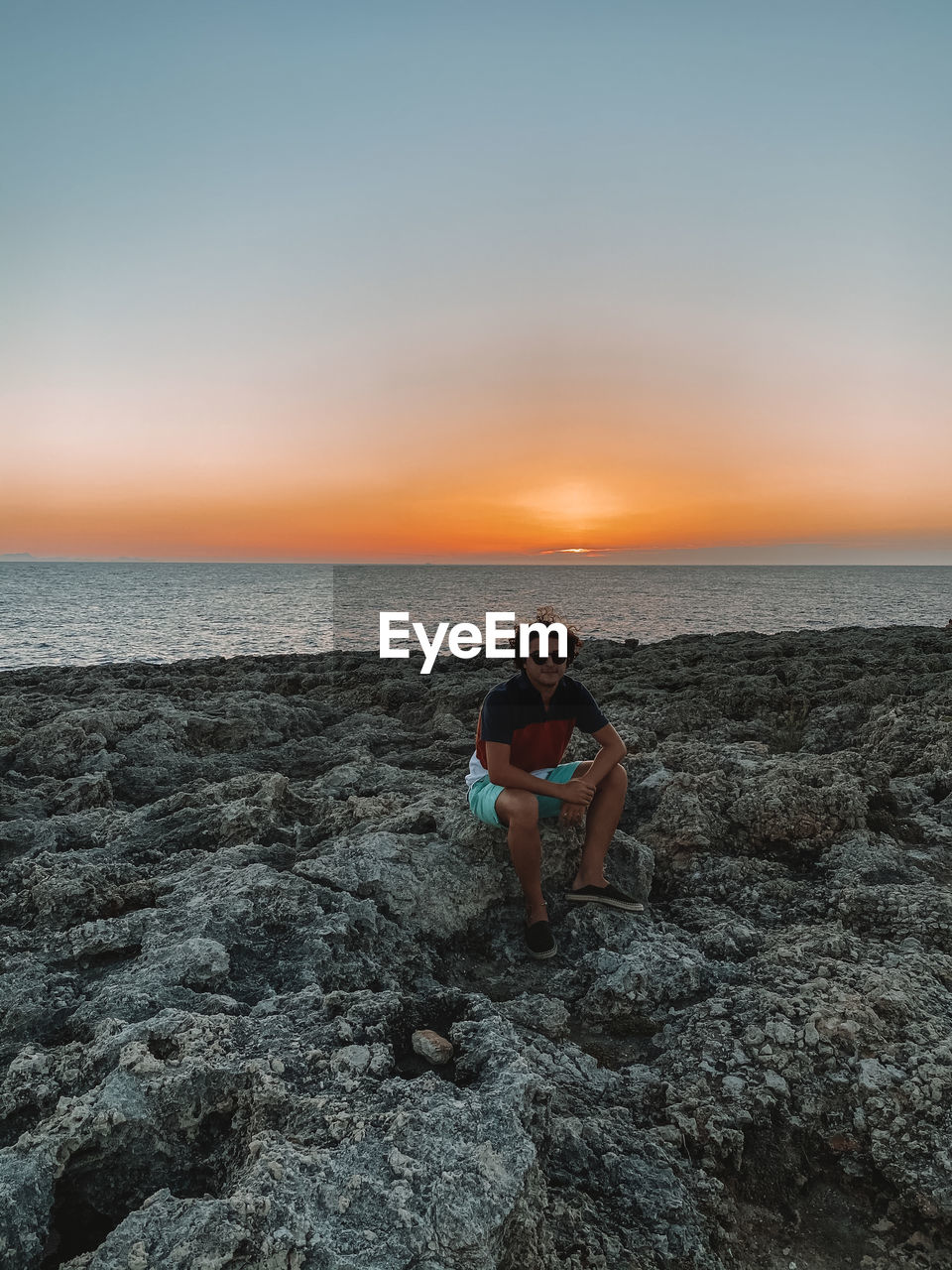 Man sitting on rock at beach against sky during sunset