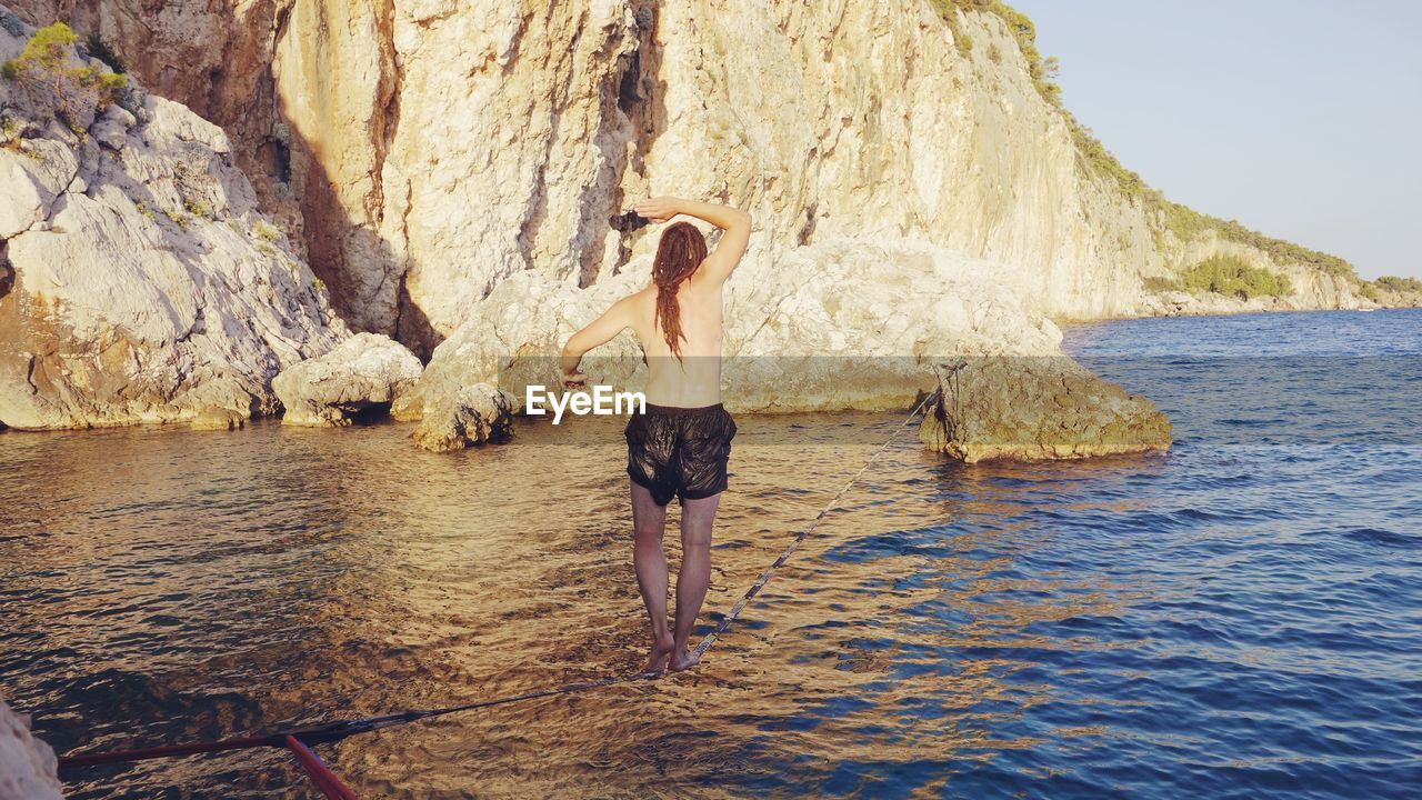 Rear view of shirtless man slacklining at sea against rocky mountain