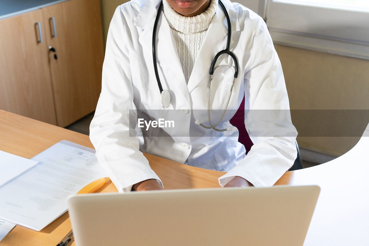 From above crop african american female doctor typing report on laptop while working at table in modern clinic office