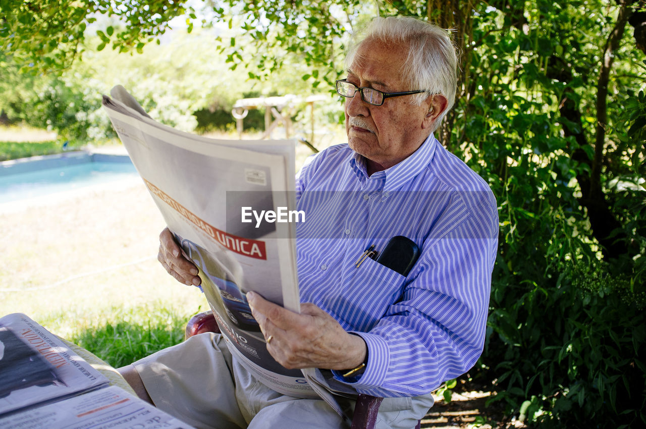Senior man reading newspaper while sitting against tree