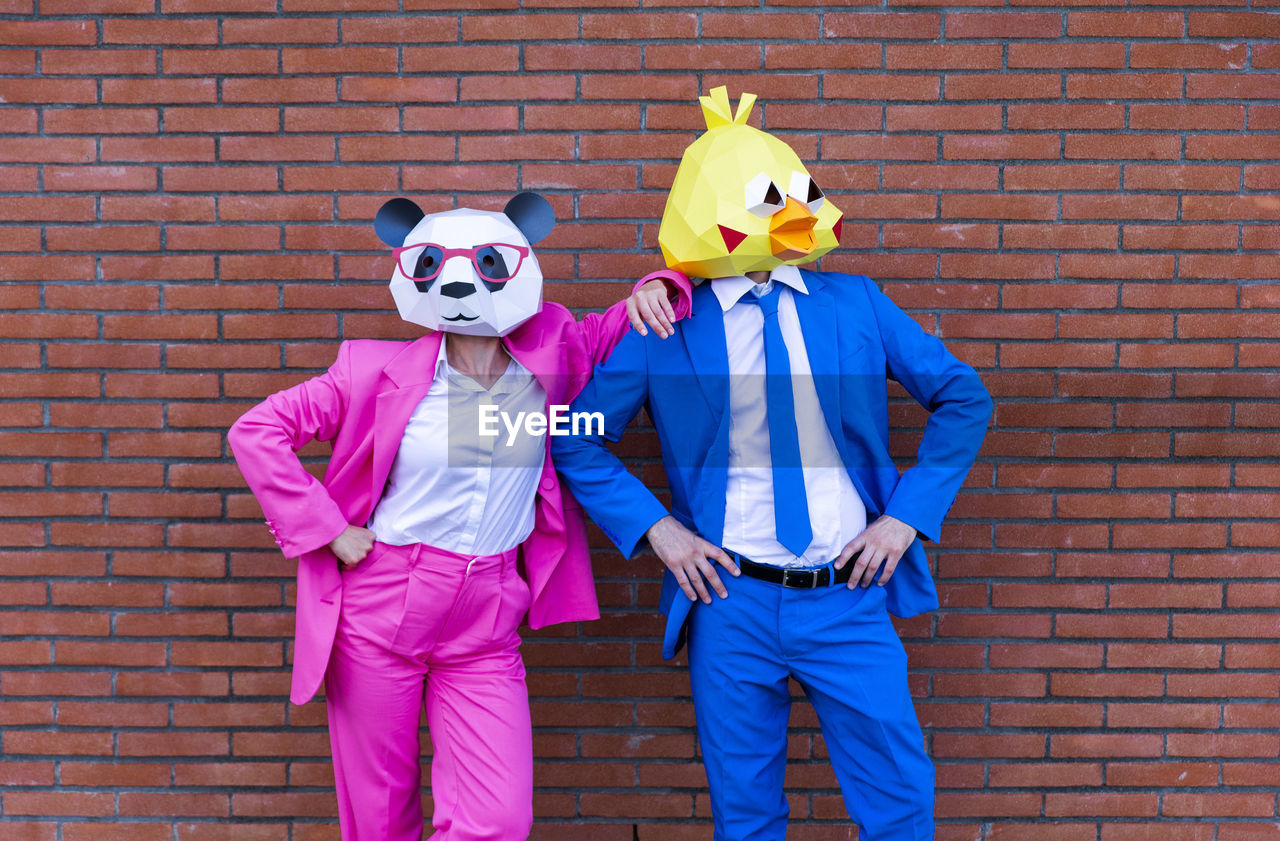 Man and woman wearing vibrant suits and animal masks posing together in front of brick wall