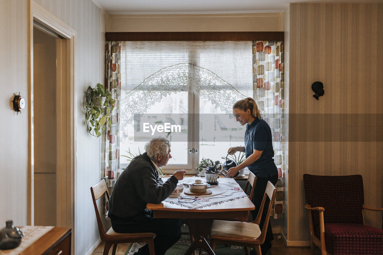 Senior man talking to female caregiver watering plants at home