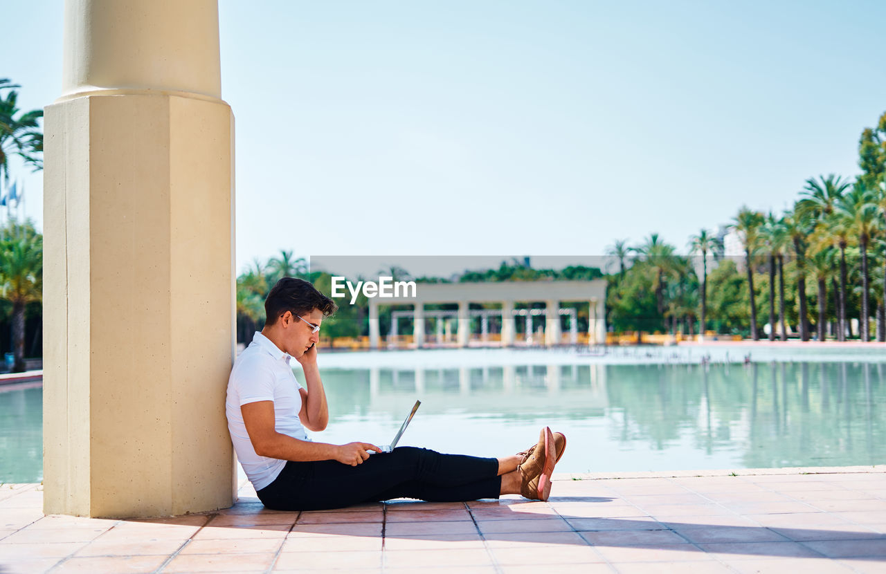 Side view of concentrated male student browsing netbook and speaking on mobile phone while sitting near stone column in university campus