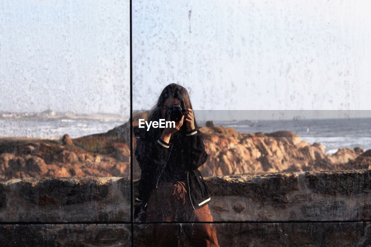 WOMAN PHOTOGRAPHING SEA THROUGH CAMERA WHILE STANDING BY WATER