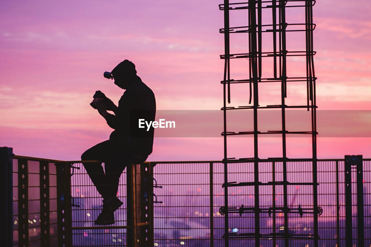 Side view of silhouette man holding camera while sitting on railing at sunset