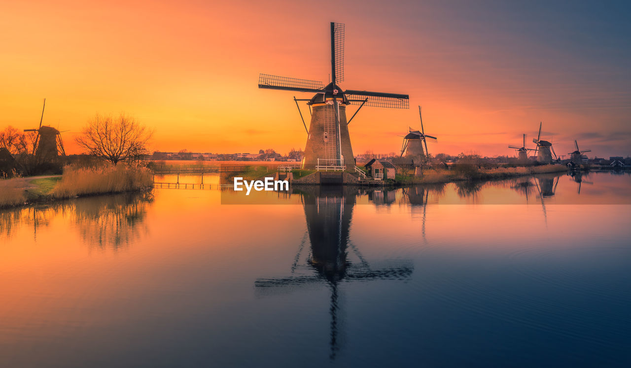 Traditional windmill by lake against sky during sunset