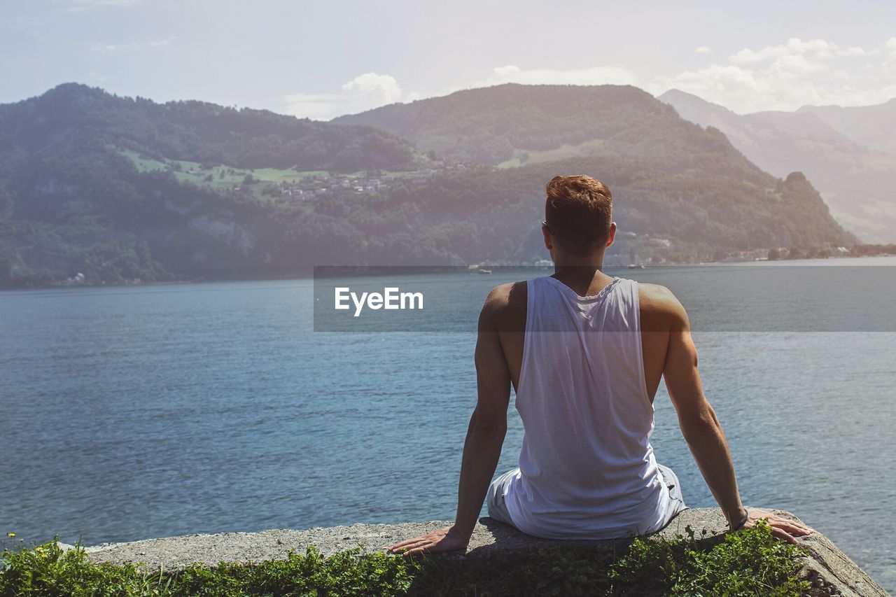 Rear view of man sitting while looking at sea and mountains