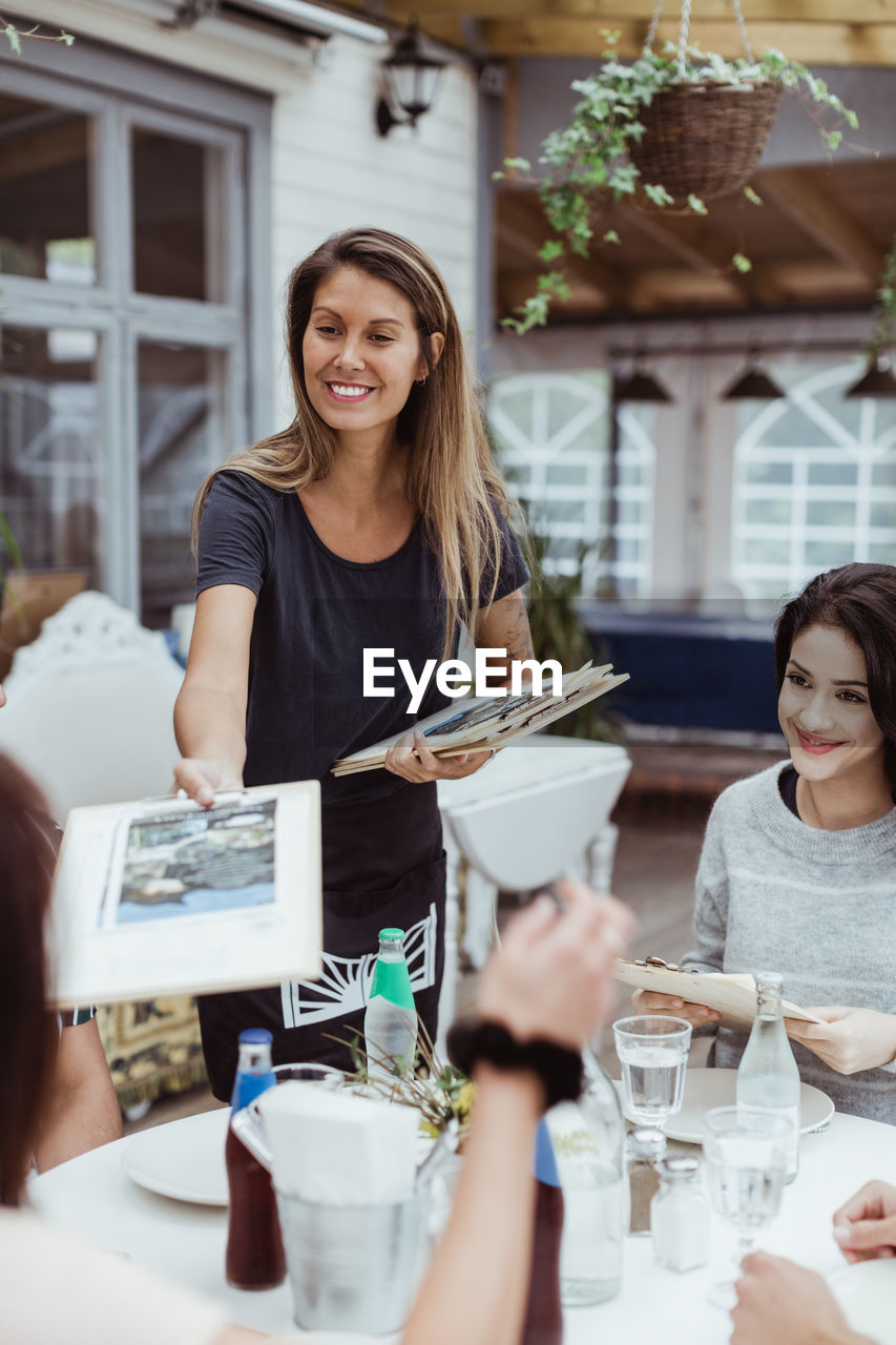 Smiling female owner giving menu cards to customer sitting in restaurant