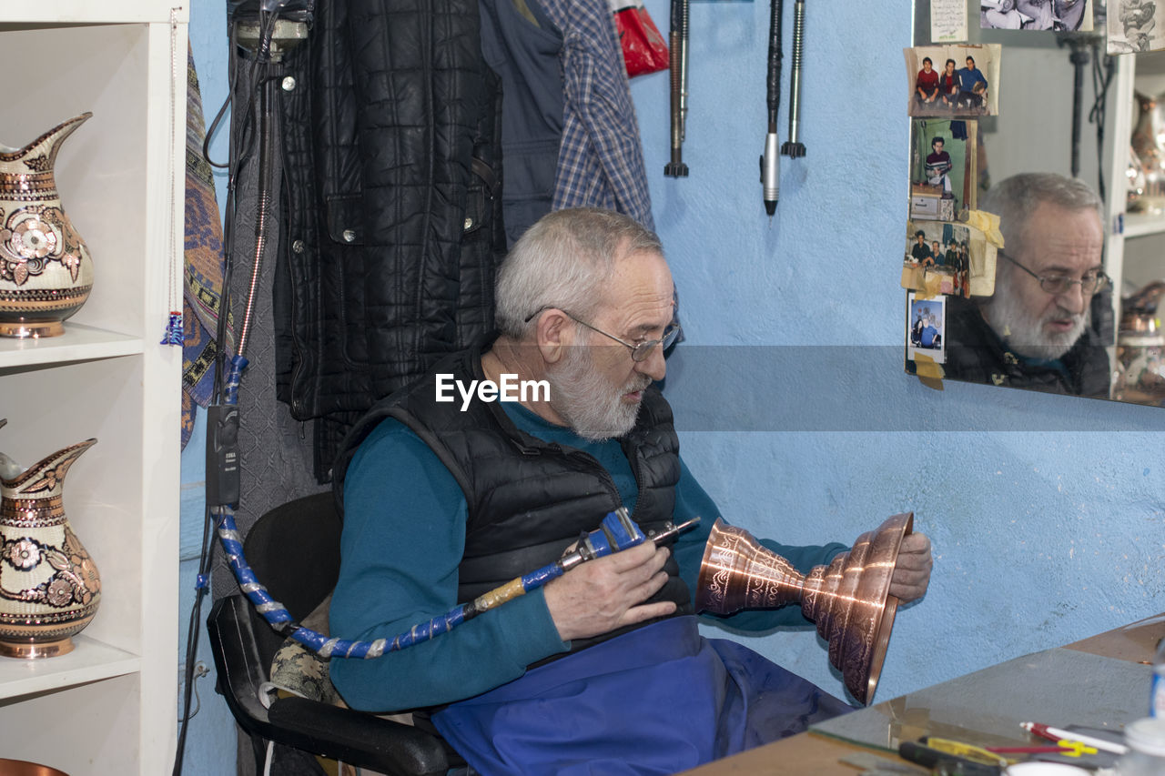 Senior man carving on brass container while sitting in workshop