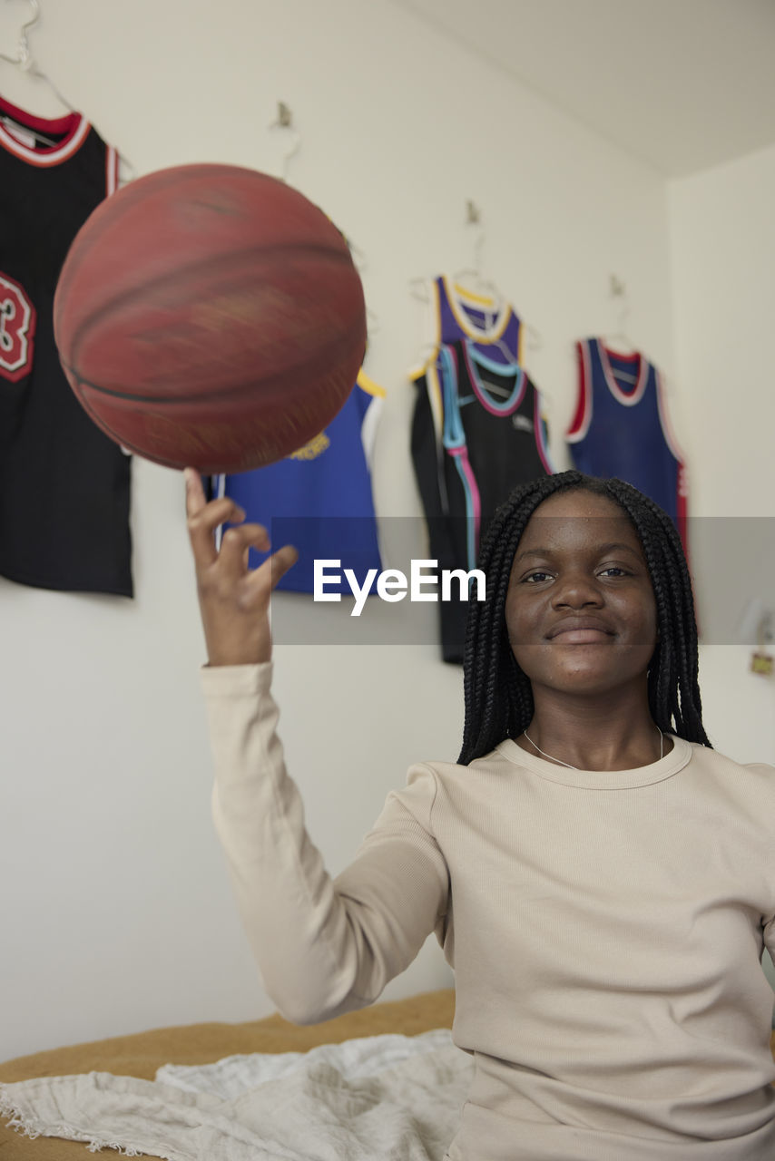 Portrait of teenage girl spinning basketball on her finger while sitting in bedroom