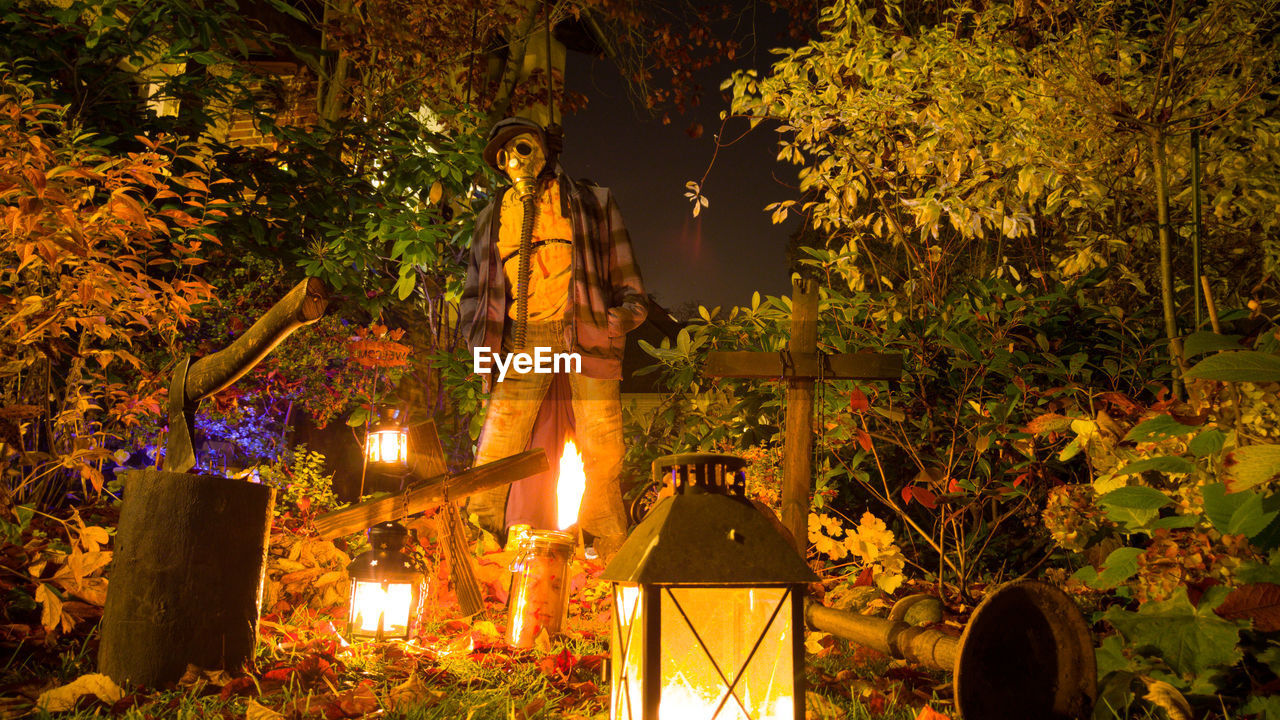 Scarecrow with gas mask amidst trees and illuminated lanterns at night