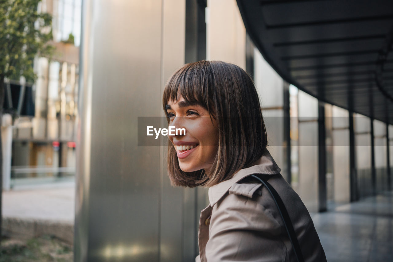 Side view of positive young female in stylish coat leaning on railing near columns while standing on terrace of contemporary building