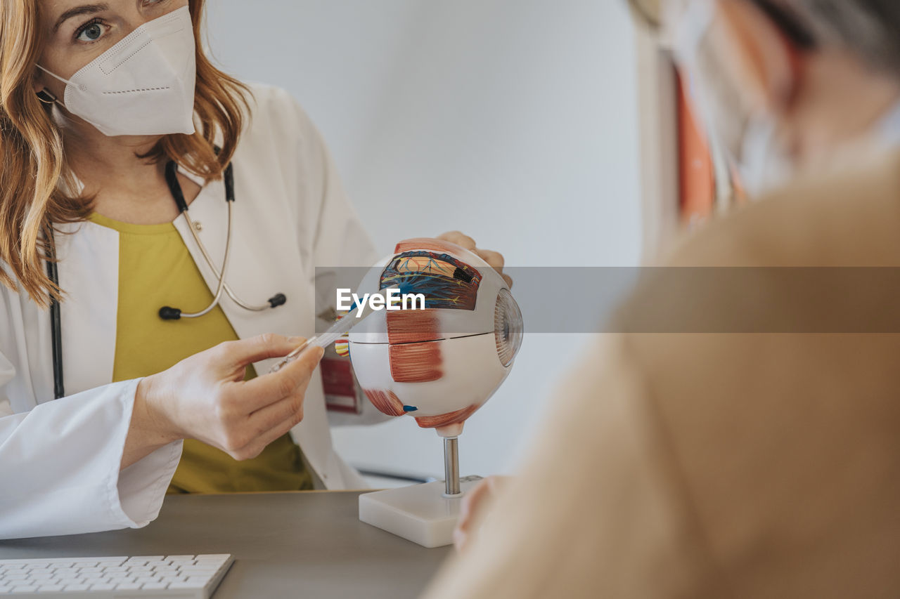 Female doctor with protective face mask explaining over artificial eye to patient at clinic