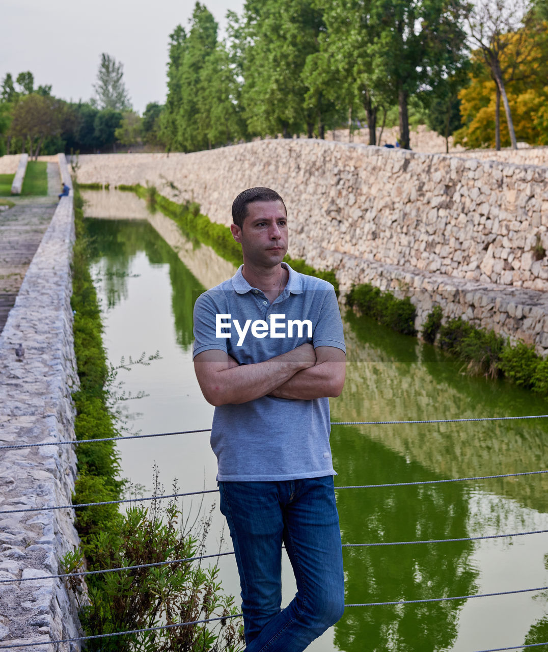 one person, water, men, adult, standing, plant, nature, casual clothing, pond, day, front view, looking at camera, portrait, full length, tree, three quarter length, leisure activity, lifestyles, river, arms crossed, young adult, outdoors, smiling, reflection, clothing, emotion, looking, occupation, architecture