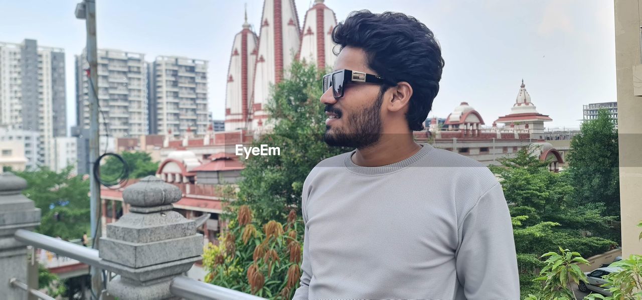 architecture, city, one person, building exterior, built structure, adult, young adult, beard, men, glasses, facial hair, casual clothing, building, nature, city life, lifestyles, standing, waist up, fashion, day, sunglasses, looking, skyscraper, portrait, sky, outdoors, leisure activity, eyeglasses, black hair, cityscape, looking away, person, tourism, t-shirt, smiling, tree, travel destinations, vacation, residential district, contemplation, clothing, cool attitude