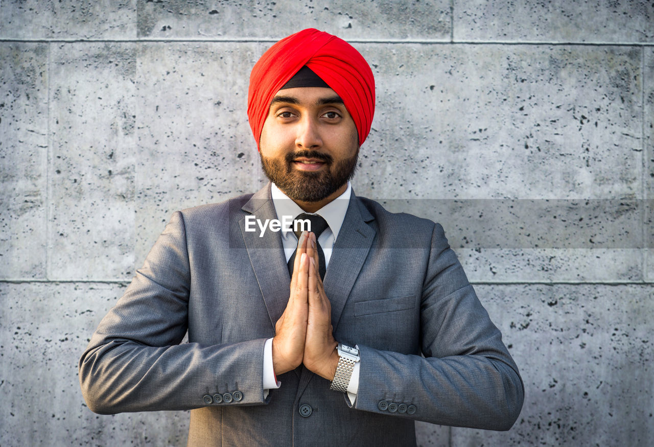 Portrait of smiling businessman wearing turban with clasped hands against wall