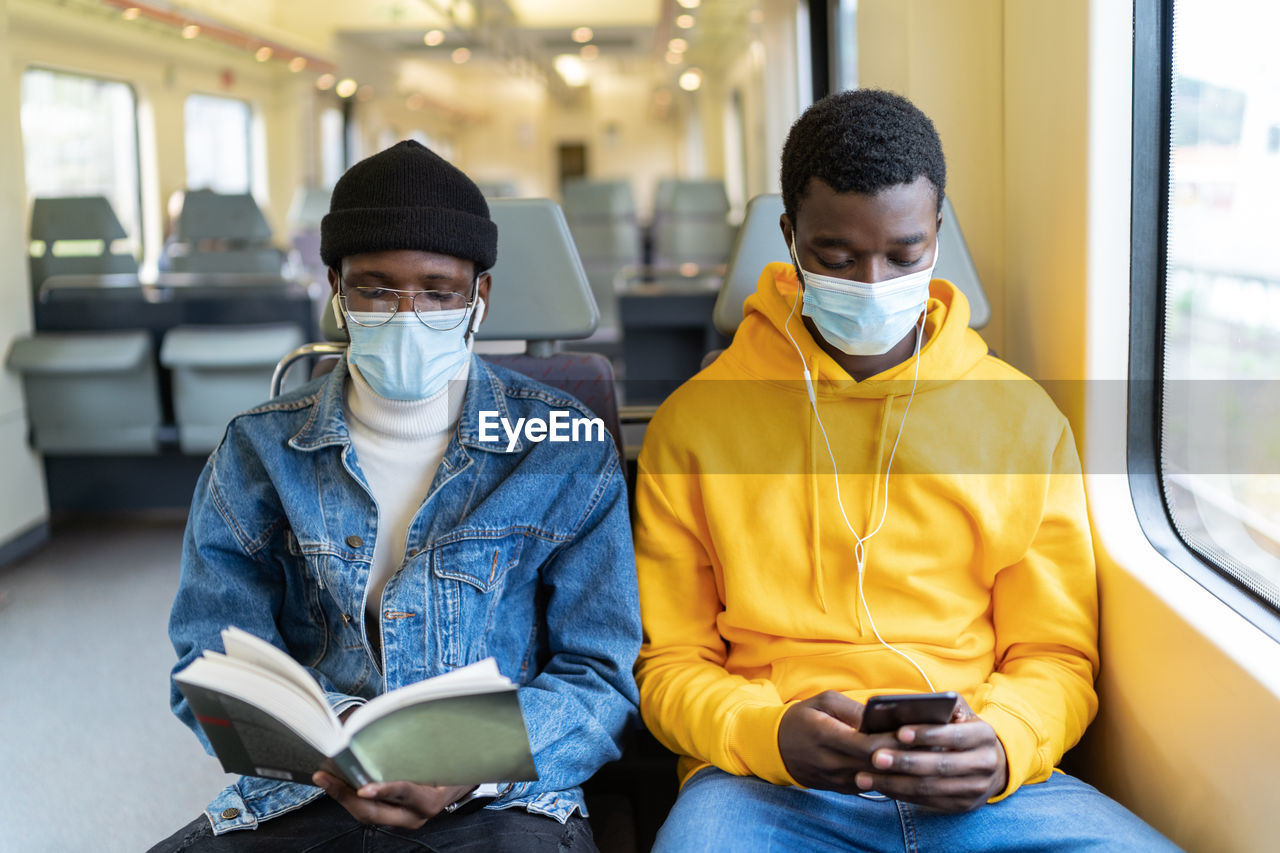 Busy african american male travelers in medical masks reading book and listening to music while sitting on passenger seats in train and entertaining during trip