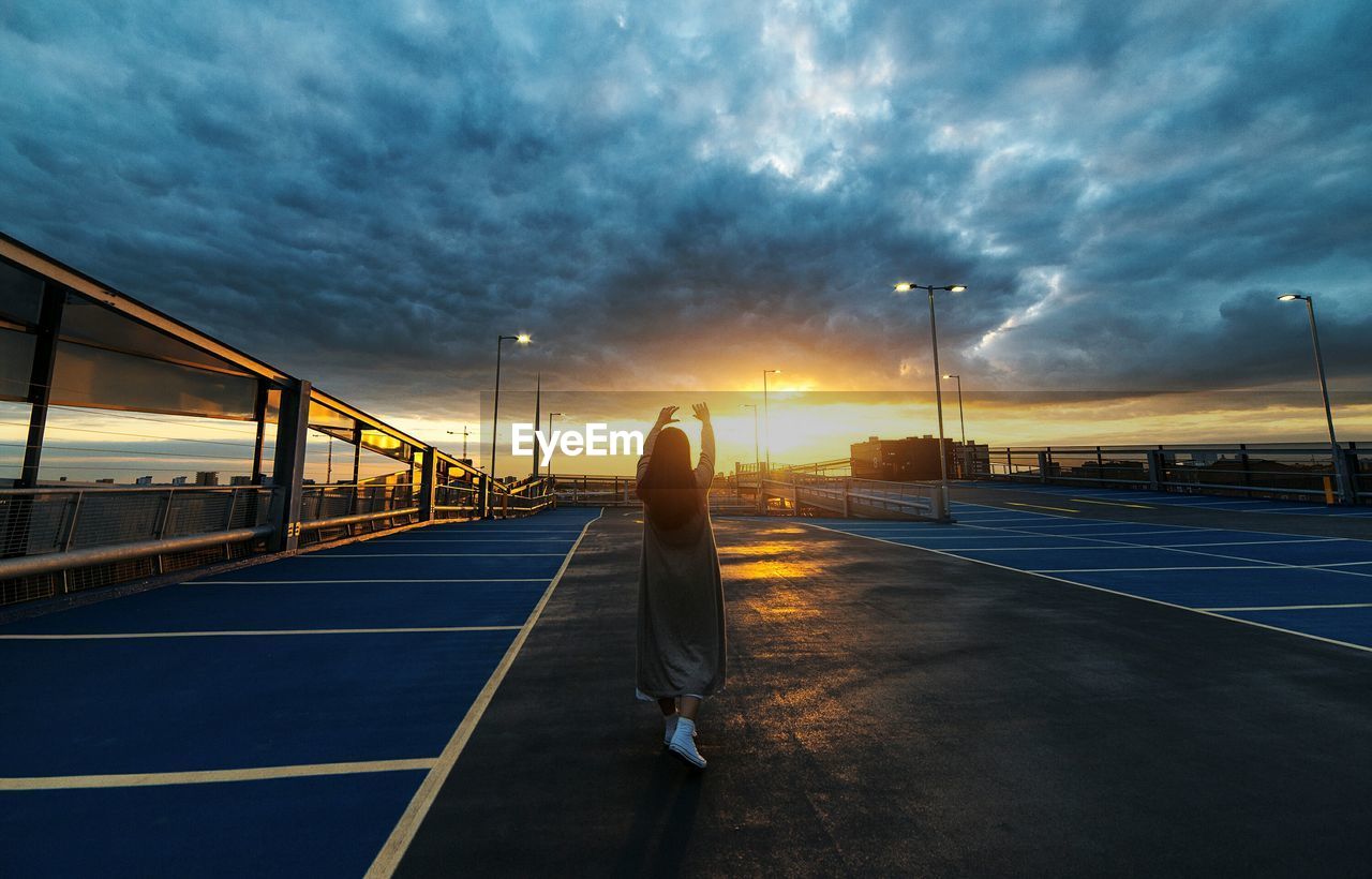 Rear view of woman with arms raised standing in parking lot against cloudy sky