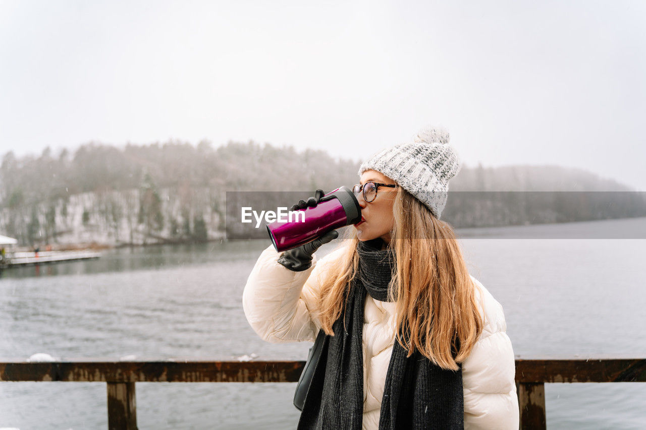 Woman drinking from a travel mug by the lake on a snowy winter day