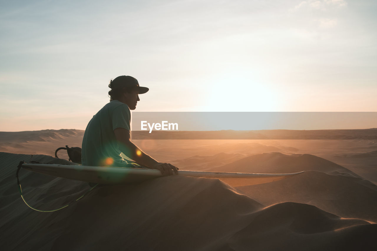 Man with sandboard sitting at desert during sunset