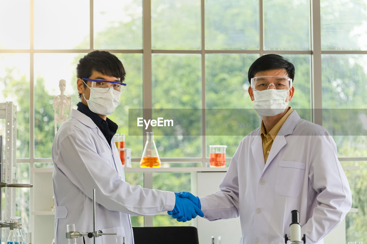 Senior and young doctor scientist wearing glasses, face mask, glove make a hand shake in laboratory.
