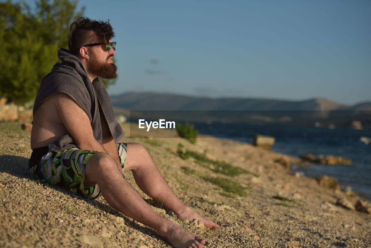 Bearded man sitting at beach against sky