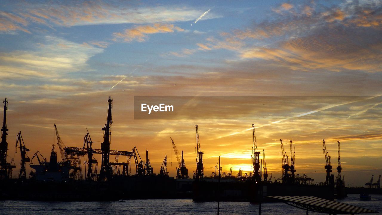 Silhouette cranes at commercial dock against sky at sunset