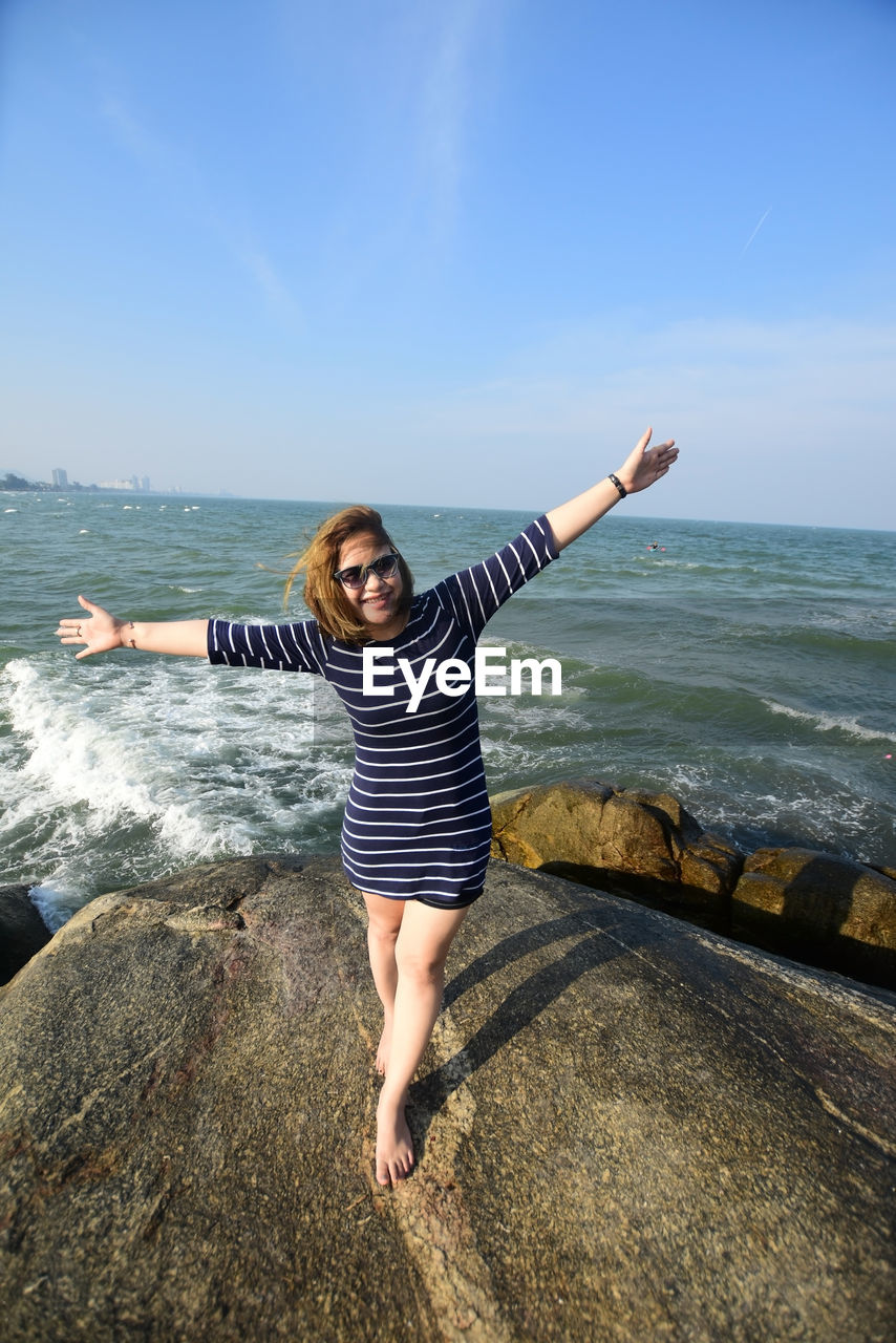 Young woman arms outstretched standing on rock at seashore against sky