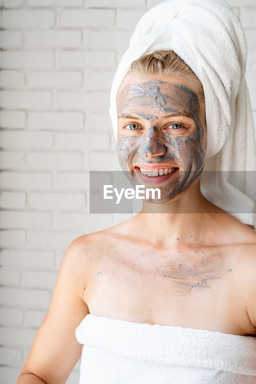 Young smiling woman wearing white bath towels with a clay facial mask on her face